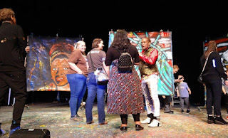 A group of 3 women speak with one of the Artrageous performers on the stage with paintings in the background.