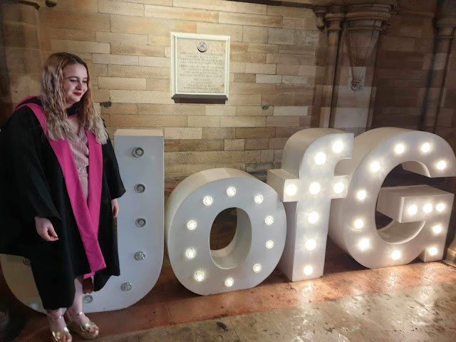 a picture of a blonde woman in a black and pink graduation robe, wearing a pink dress. She is standing in front of large, white letters: U of G.