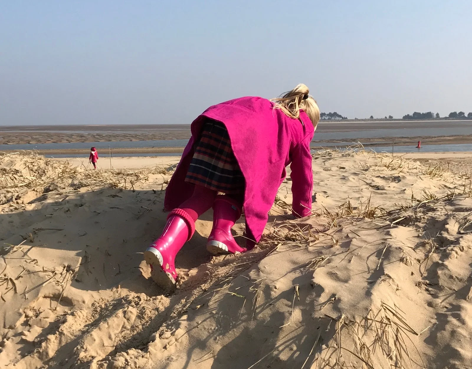 A 5 year old girl in pink coat, tights and wellies climbing up a sand dune on all fours