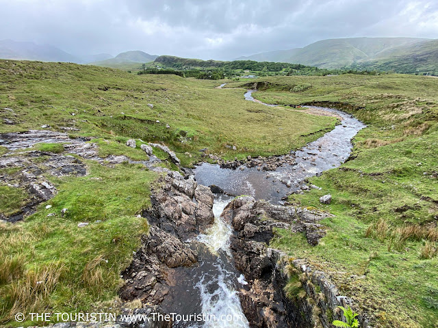 A waterfall flows into a winding stream that travels through marshland, and moorland, towards craggy mountain peaks in the far distance.