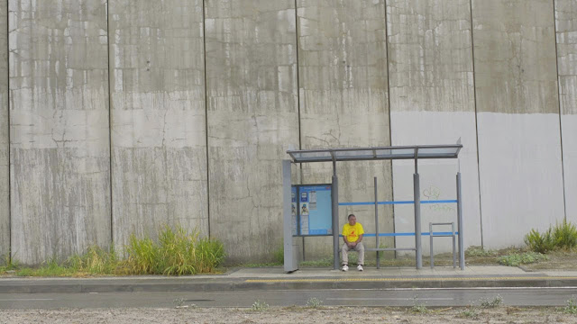 Fotograma del corto "Hombres solos", en el que se ve, desde lejos, a un hombre solitario sentando en una parada de autobús