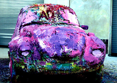 This is a classic Volkswagen Beetle covered with thick layers of finger paint.  The most dominant color is purple but there are also reds, yellow and blues.  The letters AD are carved in the paint that covers windshield.