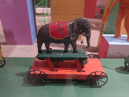 Photo of a child's pull toy. A flat red wheeled platform, with a green articulated platform on top of it. On top of the green platform is a gray elephant with a red blanket over its midsection, holding a wire with a small bell on each end.