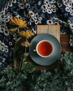 [Image Description] Top down photo of a cup of tea on a saucer on a wooden table with yellow fresh cut flowers and a black and white floral cloth, as well as an open book.