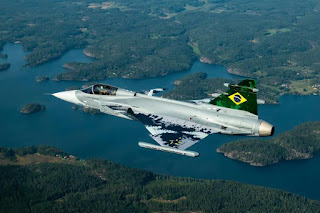 Descrição do primeiro caça supersônico Gripen E/Br, construído na Suécia para a Força Aérea Brasileira(FAB).   Foto de perfil esquerdo do caça em voo, em dia ensolarado. Com 14,1 metros de comprimento e 8,6 metros de largura, o caça multiemprego(ataque no ar,terra e mar) é uma aeronave de última geração que atinge mais de duas vezes a velocidade do som, e suporta até nove vezes a força da gravidade durante manobras. Pintado de cinza, com variações digitalizadas, a inscrição “Força Aérea Brasileira” na seção esquerda próximo ao cockpit e na cauda foi aplicada a bandeira do Brasil. O Gripen voa a 2.406km/hora e pode cobrir pouco mais de 3 mil km. Com carga máxima de ataque(um canhão de 30 mm, mais 7.2 toneladas de mísseis e bombas, seu raio de ação é de 800 km/1 mil km. O cockpit panorâmico comporta um piloto e o arrojado painel é cem por cento digital, uma tela de 19 x 8 polegadas de alta resolução. As aberturas quadrangulares das tomadas de ar localizam-se nas laterais da carenagem do caça ao longo do comprimento junto as asas e o motor turbo fan, localiza-se na parte traseira, abaixo da cauda. A asa em delta, em formato de triângulo, é projetada especificamente para que quando ela ultrapassar a barreira do som toda a asa fique dentro da onda de choque. No nariz do caça, um radome(componente muito resistente que suporta forças aerodinâmicas e eventuais birdstrikes(colisão com pássaros) e impactos com chuva e granizo, ao mesmo tempo, permite a passagem das ondas de radar e bem na ponta, o tubo de Pitot (sensor de pressão/velocidade, uma espécie de cano fino de aço com um furo na extremidade). Lá embaixo, vista panorâmica de uma represa com vegetação densa e algumas casas. 