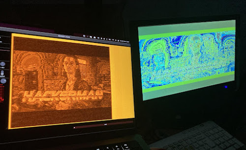 [Image: On the right, a monitor shows a noisy green and blue image. On the left, another monitor shows a grainy picture of a man and the text 'Hackerman'.]