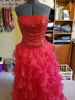 Dress is on a dress form. Sleeveless, red sequined top and lots of red layers flaring out from the waist to the floor.