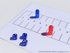 na zdjęciu plansza do gry Marinetti w kształcie prostokąta z 12 polami, na polu nr 6 stoi czerwony pionek, na polu nr 3 stoi niebieski pionek a na pierwszym planie widać trzy niebieskie kostki z wynikami  2,2,5