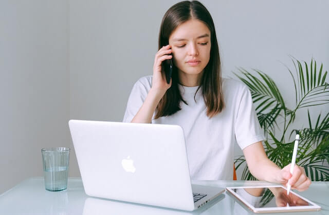 Woman in white shirt calling on her mobile, writing on iPad with laptop on the table