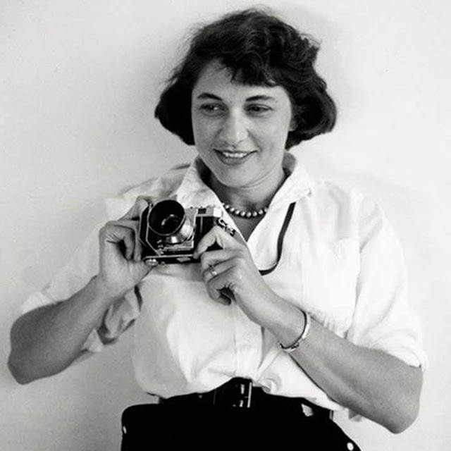 Ruth Orkin com o semblante feliz e sorriso no canto do rosto está segurando uma câmera fotográfica. Seus olhos aparentam estar direcionados para uma figura que a traz felicidade. Ela usa uma blusa branca de botão, uma calça preta e um cinto da mesma cor. Em seu pescoço, a fotógrafa carrega um cordão de pérolas; vestimenta e acessórios muito característicos da época.