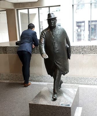 Life sized bronze statue of a portly businessman in buttoned coat and hat, holding a briefcase, captured in mid-stride