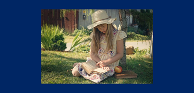 A young girl with long blond hair and strawhat and floral dress is sitting in a garden reading.