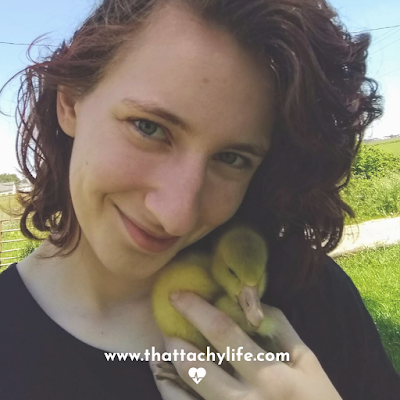 Bonje Gioja holds a fluffy baby goose up to her cheek while she smiles at the camera. Her curly red hair is cut to shoulder-length and blowing in the wind. The baby goose is craning his neck around to get a look at the camera too.