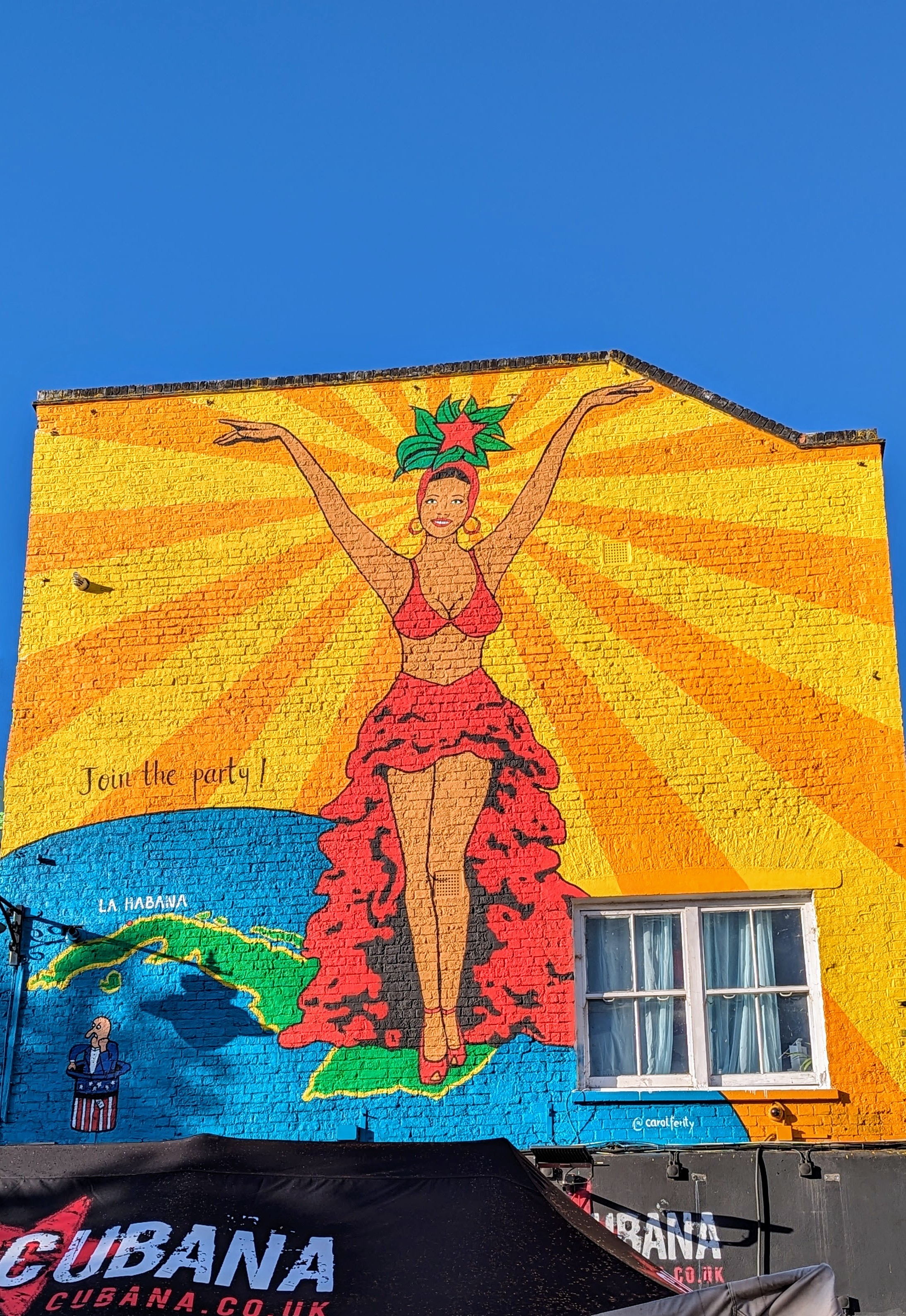 Whole wall mural of a Cuban woman dancer, red frilly skirt, red high heel shoes, and a hat if flowers stands with arms in the air against stylised sun rays and bottom right is a map of Cuba. The real sky is blue as!