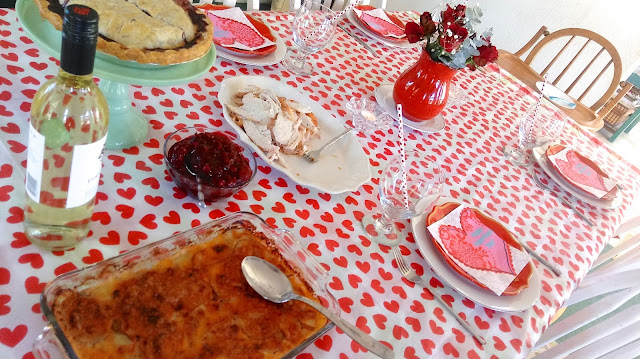 a table covered with a heart table cloth with a red vase of roses int he center, a blueberry pie on a cake stand, a scalloped potato casserole, a platter of roast turkey, a bottle of wine, and pretty table settings for each person