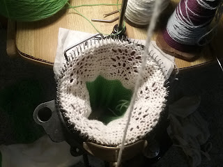 A circular sock knitting machine with a lacy piece of knitting in progress.