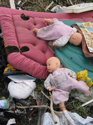 Close up picture of a rubbish heap, features burnt headboards and two baby-like dolls one lying face-down, one face-up