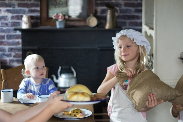 A girl dressed up in a mop cap and apron, a sack of flour, pretend bread and a kettle