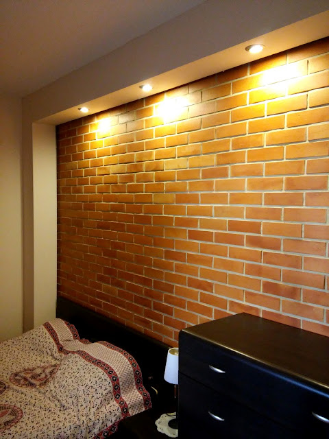 Ceglana ściana w sypialni. Barwa cegieł wyeksponowana jest dzięki kinkietom w podwieszanym suficie.