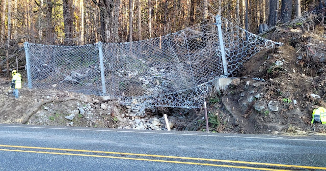 Work crews placing a tan erosion blanket over the earth berm along US 2. A dump truck towing a trailer with construction equipment sits along the shoulder of the highway.