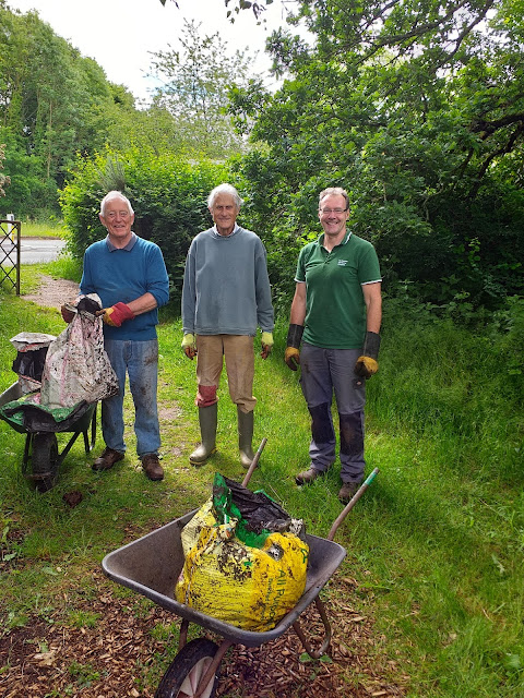 three men stood wearing gardening gloves and welly boots beside a bag of manure in a wheelbarrow