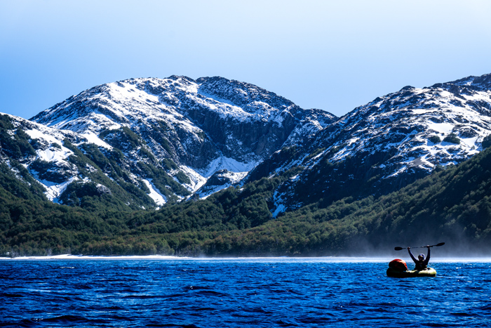 A paddler is on the lake, holding up his paddle. In the background is a beach.