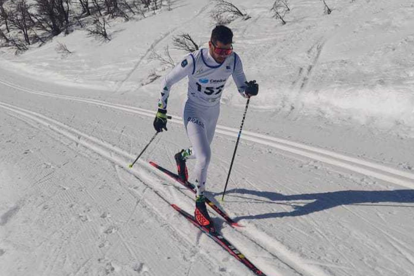 Victor Santos, com roupa e colete brancos e com detalhes em azul, utiliza bastões para andar na neve e representar o esqui cross-country do Brasil