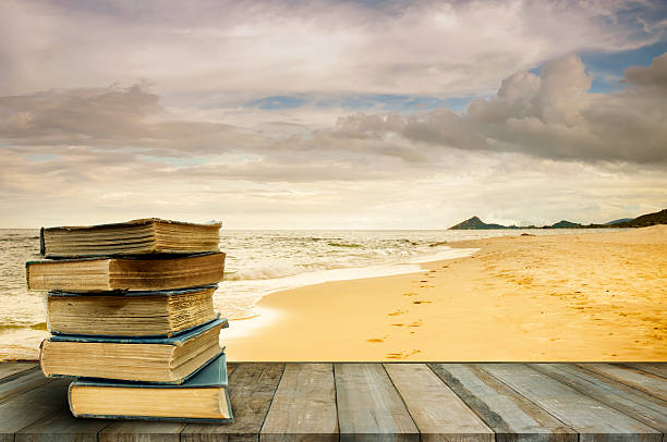 a stack of weathered hardback books on a wooden deck in front of a sandy beach