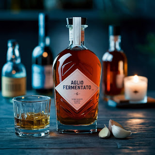 "Whiskey bottle labeled Aglio Fermentato, sliced garlic, wooden surface, blurred background, additional bottles, candle, cinematic shot, medium shot, whiskey and garlic, rustic setting, warm ambiance, atmospheric photography."