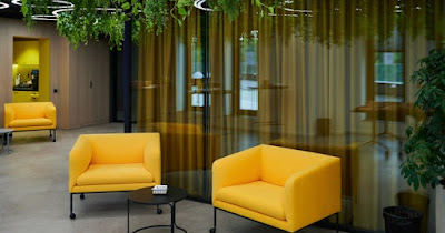 An interior view of a modern office building that has yellow chairs, glass walls, and plants hanging from the ceiling.