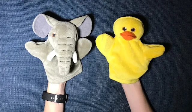 A small elephant hand puppet and a bright yellow duck puppet on the end of arms on a blue fabric background