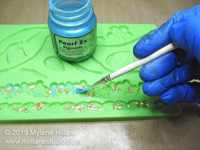 Using a soft paint brush to dust Blue-Green Pearl Ex powder around the gold leaf in the mould.