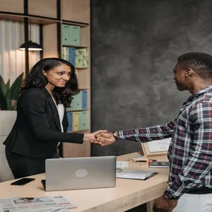 Foto de dois profissionias se cumprimentando, uma mulher de frente e um homem de lado, com uma mesa de escritório entre ambos.