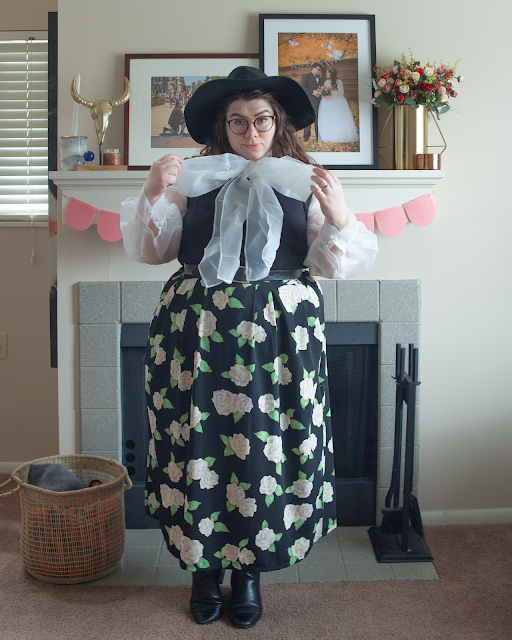 An outfit consisting of a wide brim black hat, a white organza blouse with a large bow tie under a black sleeveless dress that is tucked under a white and green floral on black maxi skirt and black chelsea boots. 