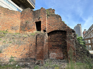 A photo of the ruins of Bastion 14 that face out towards the road.  It shows a ruin of red bricks with an arched area on the right hand side and a doorway near to the top of the building.  The sky above is blue and in the distance can be seen a grey concrete London tower block.  Photo by Kevin Nosferatu for the Skulferatu Project.