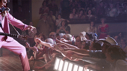 A gif from the film ELVIS. Austin Butler playing Elvis is singing on stage. He bends one knee, leans down towards the front row of screaming fans, and touches their hands as he continues to perform.