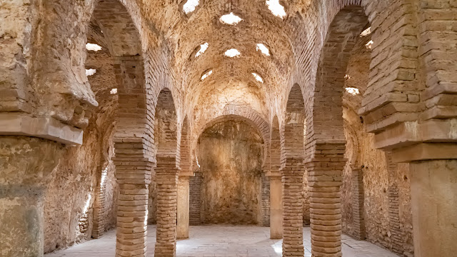 Interior de antiguos baños árabes con columnas y arcos de piedra, y tragaluces de estrella en el techo.