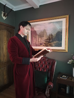 John M. Gilheany wearing a long burgundy smoking jacket in a decadent scene which includes a chesterfield chair in a matching colour, a large painting with a gilded frame and antlers (shed not shot at) on a plaque above a dark stained panel door