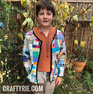 A young lad standing in the garden wearing a colourful patchwork quilt coat with a black border trim.