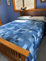 A denim quilt on a bed. A calico cat is sitting in a window overlooking the bed.