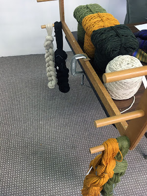 Three-quarter shot of the end of a dark wood table, with a pale wood frame clamped to it. Thick crocheted chains of wool yarn hang from the lowest pegs of the wooden frame; white and dark green on the far peg, and marigold and light green on the near peg. Matching cakes of wool yarn sit on the table, just to the right of the clamped frame, with deflated cakes of light green, marigold, and dark green sitting on top of their matching full cakes.