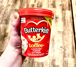 A red cylindrical tub filled with ice cream with a circular red lid with some golden brown popcorn with butterkist toffee and popcorn flavoured ice cream in gold font on a bright background