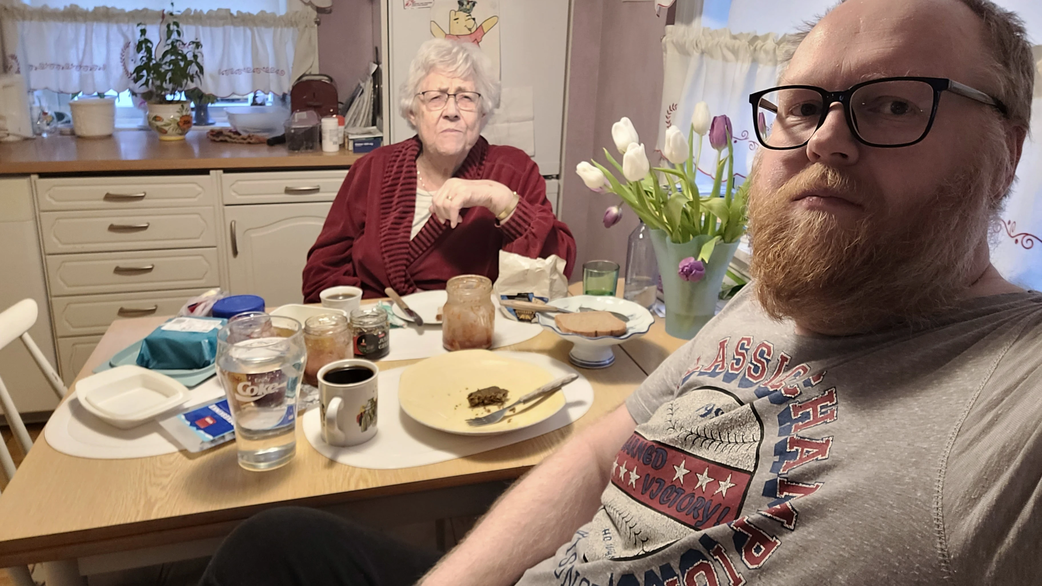 En eldre kvinne med hvitt hår og briller sitter ved et kjøkkenbord iført en rød morgenkåpe. Foran henne står brød, syltetøy og kaffekopp. Ved siden av sitter en mann med skjegg og briller, iført en grå t-skjorte. På bordet står også en vase med tulipaner. De sitter sammen til frokost på et kjøkken med hvite skap og gardiner med blondekanter.