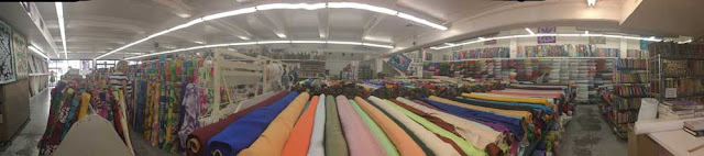 A panoramic photo of the inside of a massive fabric shop with bolts and rolls of brightly colored fabrics stacked and propped everywhere. The fluorescent ceiling lights have been stretched amusingly by the panorama.