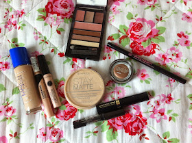 selection of makeup on floral duvet: beige foundation bottle, two beige tubes, beige powder, brown shimmery tub, three black tubes and open palette with browns and pinks