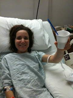 I'm in a hospital bed, wearing a hospital gown, holding up a drink in a Styrofoam cup.