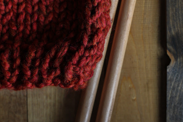 Bottom quadrant of a finished rust chunky yarn hat sitting on a wooden surface next to large bamboo knitting needles.