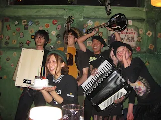 Group of musicians standing in front of a green wall covered in coassters from Irish pubs. Band is 5 people in the back holding guitar, mandolin, cajon, and accordian with one band member in the front honding a birthday cake