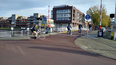 Une piste cyclable rouge traverse un pont aux balustrades blanches. Il y a des gens qui traversent le pont à vélo. Il y a aussi des barrières rouges et blanches qui peuvent fermer le pont.
