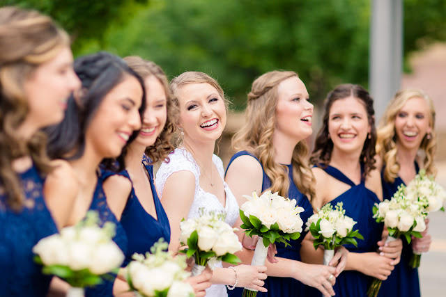 On the morning of Saturday, July 8th, the rainclouds were threatening to unleash their fury, but it didn’t matter because this was Anna and Ben’s wedding day, and for that reason alone, this day would sparkle! From the moment we got to the church, activities were in full gear…fun and impromptu pictures of the bride with her bridesmaids, as they assisted Anna  into a wedding dress that was seemingly made for her…a mermaid-shaped, intricately laced, floor length, white  gown, with a scalloped V-neck front, and a diamond-shaped, cut-out in the back.  An embroidered handkerchief was tucked inside her garter as a “remembrance” of her grandmother.  The six bridesmaids wore individually designed, royal blue chiffon gowns, and each carried a similar version of Anna’s lush bouquet of pale, ivory roses and white lilies.    Impromptu shots of the relaxed and charismatic groom with his groomsmen came next.  The comradery they share was evident in every photograph!  Then it was time to squeeze in a sentimental “first look” for the bride’s dad before the romantic “first look” with the groom, and you can tell from the expression on Ben’s face…and the height of his jump…how excited he was to finally see her!     First Church OKC was an ideal location for this couple’s wedding.  Located in the heart of Oklahoma City, its ministry began during the land run of 1889, and has continued to share the gospel through two wars and the infamous April 1995 bombing.  The large, stained glass windows, dark mahogany wood, and simple, yet elegant sanctuary was the perfect backdrop for these two young people who were meant for each other.     When asked what the best part of the day was, Anna said, “getting to see Ben for the first look, and his reaction to seeing me.”  Ben’s response was “getting to see Anna walk down the aisle and say our vows to each other.”     Anna felt that her wedding day went smoothly, but if she were to do it over again, she would hire a wedding planner and a florist to take some of the responsibility off herself, family, and friends.  Her favorite piece of advice to a prospective bride and groom:  “When things get stressful or overwhelming—and at some point they will—just take a break, step back, and enjoy.”    After the couple exchanged vows, guests were invited to participate in a fun, romantic, and lively reception that included the “first dance,” dances with the parents, toasts to the bride and groom, a sit-down dinner, the cake cutting, throwing of the bouquet and garter…and lots and lots of dancing!  At the end of the evening, it was time for friends and family to gather together and wish this fun-loving couple farewell and best wishes for a wonderful life together!  